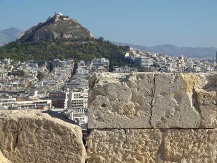 Close-up of an ancient stone wall, behind it a city and a hill with green vegetation, ancient