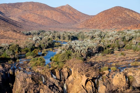 Kunene River with green vegetation in dry red mountain landscape, waterfall and African Baobab