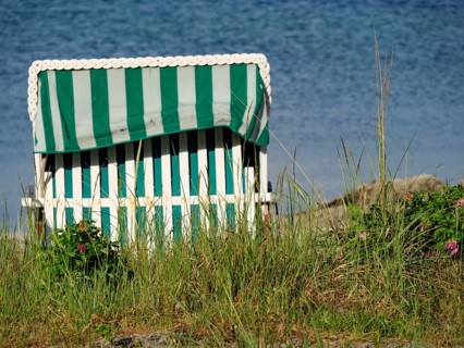 A green striped beach chair on the beach, surrounded by grass and flowers with the sea in the