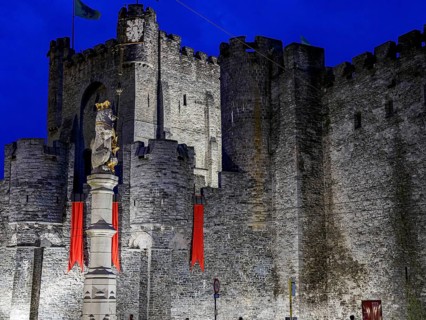 Medieval castle with massive stone walls, a statue and red flags illuminated at night, historic