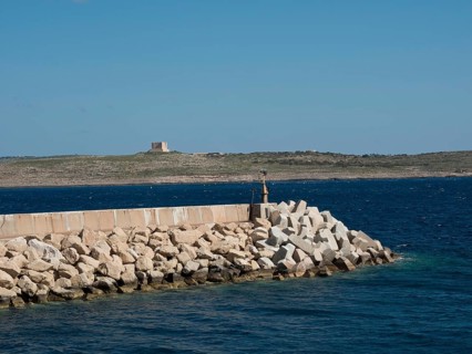 A breakwater of large stones in the sea with a rocky coastline in the background, the island of