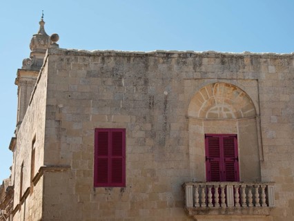 An old building with red shutters and a small balcony under a clear sky, the town of mdina on the
