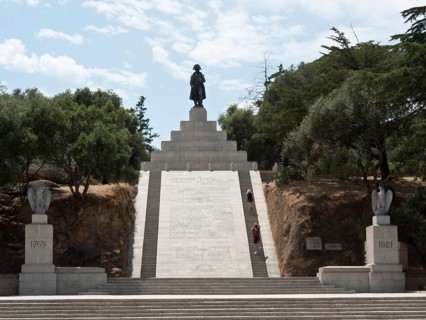 Large monument with a statue on steps, surrounded by trees and a clear sky in the background,