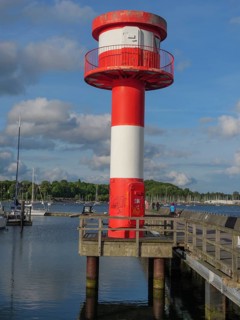 Close-up of a red and white lighthouse on a rough wooden jetty with blue sky and clouds, harbour