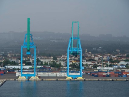 Two large blue harbour cranes in front of a cloudy industrial landscape, several containers in the