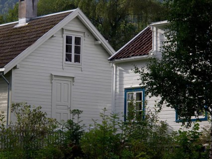 Two white wooden houses with windows, surrounded by a garden and trees in green surroundings, grey