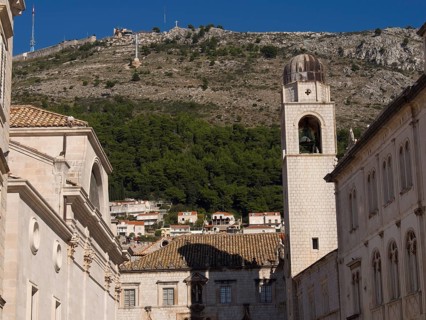 Church bell tower and historic buildings in front of a wooded mountain and blue sky, typical for