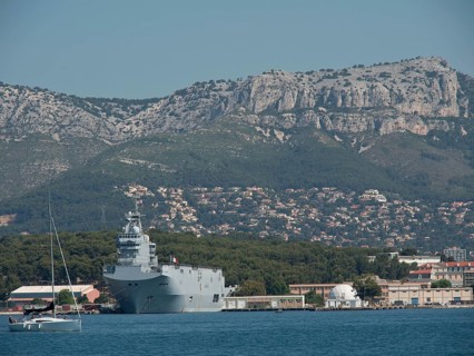 A warship lies next to a sailing boat in the harbour, with a mountain range in the background and a