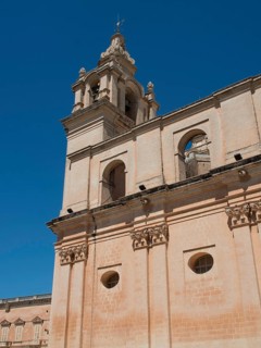 A tall church tower and sand-coloured buildings under a clear blue sky, the town of mdina on the