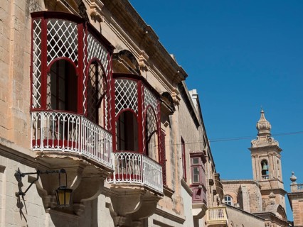 A view of an old town street with several balconies and a church tower in the background,