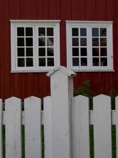 Red wooden house with two large white windows behind a willow fence, colourful wooden houses behind