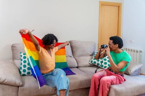 Gay man posing for his couple taking photos raising LGBT rainbow flag at home