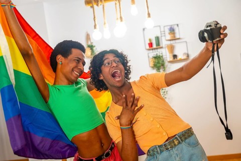 Gay latin young couple taking a selfie with a camera while raising rainbow lgbt flag at home