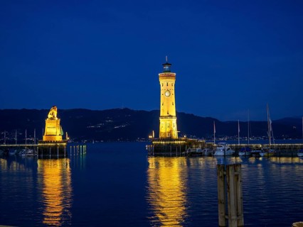 Illuminated lighthouse and lion statue at night, whose lights are reflected in the water of the