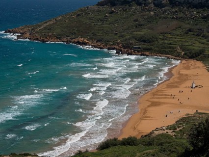 View of a quiet beach with golden sand and waves in the turquoise sea, the island of Gozo with