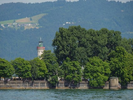 A lighthouse behind trees on the shore of a lake, in the background mountains with forest, view of