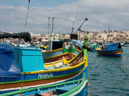 Several fishing boats in the harbour of a town, traditionally painted under a blue sky, many