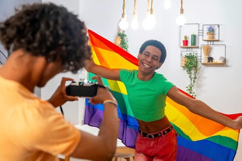 Gay man taking photos while couple posing waving LGBT rainbow flag at home