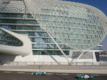 Two racing cars drive alongside a modern building with a lattice structure on the race track in