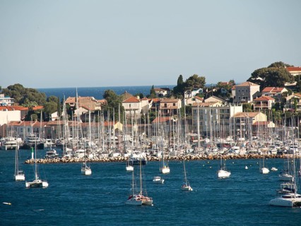 Overview of a marina with many sailing boats lying in front of a coastal town and a blue sky, la