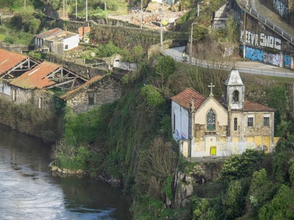 Small chapel and surrounding buildings on an overgrown, rocky cliff by a river with graffiti on