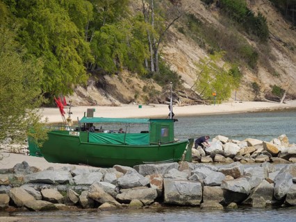 A green boat lies on a rocky shore, in the background dense forest and sandy beach, spring on the