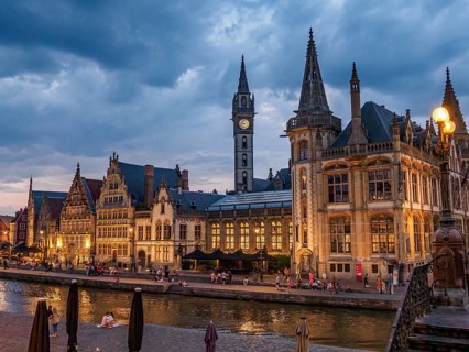 Gothic buildings with illuminated windows and people walking along a canal under a cloudy sky,