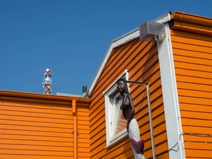 An orange-coloured house wall with a statue of a mermaid in front of a clear blue sky, Heligoland,