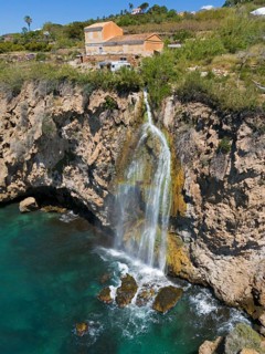A picturesque waterfall falls from a cliff into turquoise water with houses in the background, a