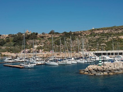 Marina with many sailing boats and adjacent hills under a bright blue sky, the island of Gozo with
