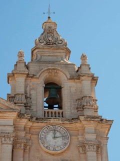Bell tower of a baroque church with clock under a clear sky, the town of mdina on the island of