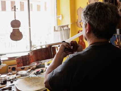 From behind, a luthier works intently on a cello in a well-organized workshop with natural light