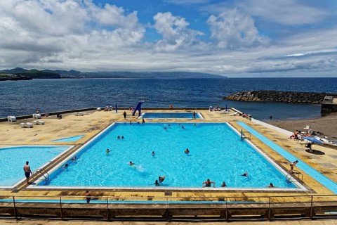 Large seawater swimming pool, filled with people, under a sunny, partly cloudy sky, coastal town of