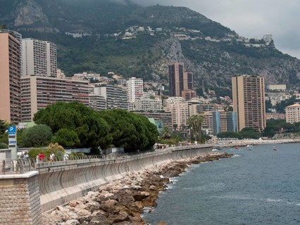 Coastline with promenade, skyscrapers and mountains behind, surrounded by sea and cloudy sky,