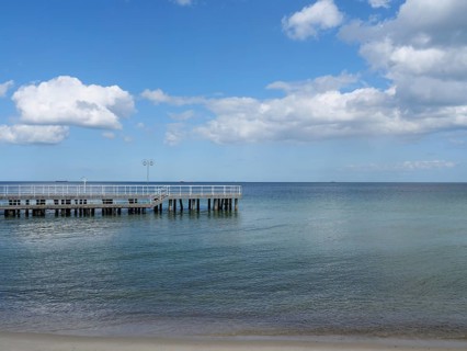 A jetty juts out over the calm waters of the sea, the sandy beach leads into the distance under a