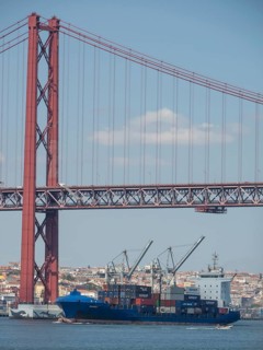 Red suspension bridge over the water, below container harbour with large cargo ships, Lisbon,
