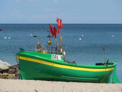 A green boat with red flags stands on the beach, propped up on the sand, in front of a calm sea,