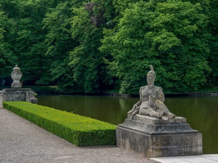 Stone statue in a park next to a lake, surrounded by dense green trees, historic castle with a