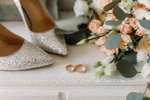 Elegant glitter shoes and wedding rings placed near a wedding bouquet of white flowers and peach