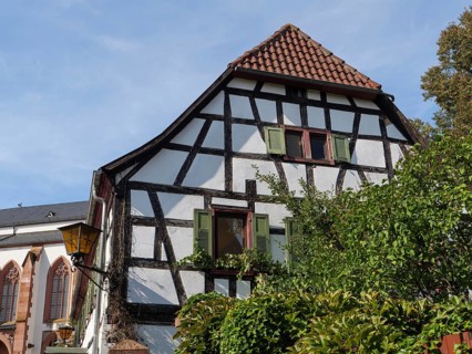 A half-timbered house with green shutters and garden plants next to a gothic church under a sunny