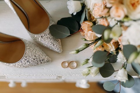 Silver shoes and wedding rings on a white table next to a bouquet of flowers