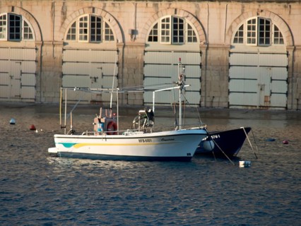 A boat in the harbour in front of a building with stone walls and many windows, small boats in the