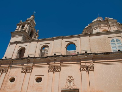 Baroque church facade with decorative elements and two towers against a blue sky, the town of mdina