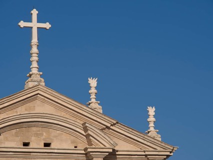 Top of a church with a cross and classical style elements in front of a cloudless blue sky, the old