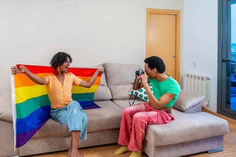 Gay man holding LGBT rainbow flag while boyfriend taking photos sitting on the sofa at home