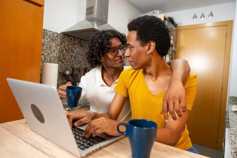 Romantic latin gay couple using laptop and drinking coffee sitting in a domestic kitchen at home