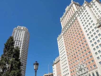 Two tall, modern buildings under a clear blue sky, a street lamp and a tree in the foreground,