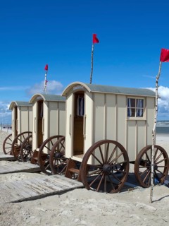 Three beige changing cabins with large wooden wheels on a beach under a blue sky with clouds,