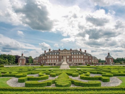 A large historic castle surrounded by a symmetrical garden with manicured hedges under a cloudy