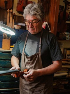A luthier wearing glasses and an apron is intently working on a cello under a desk lamp